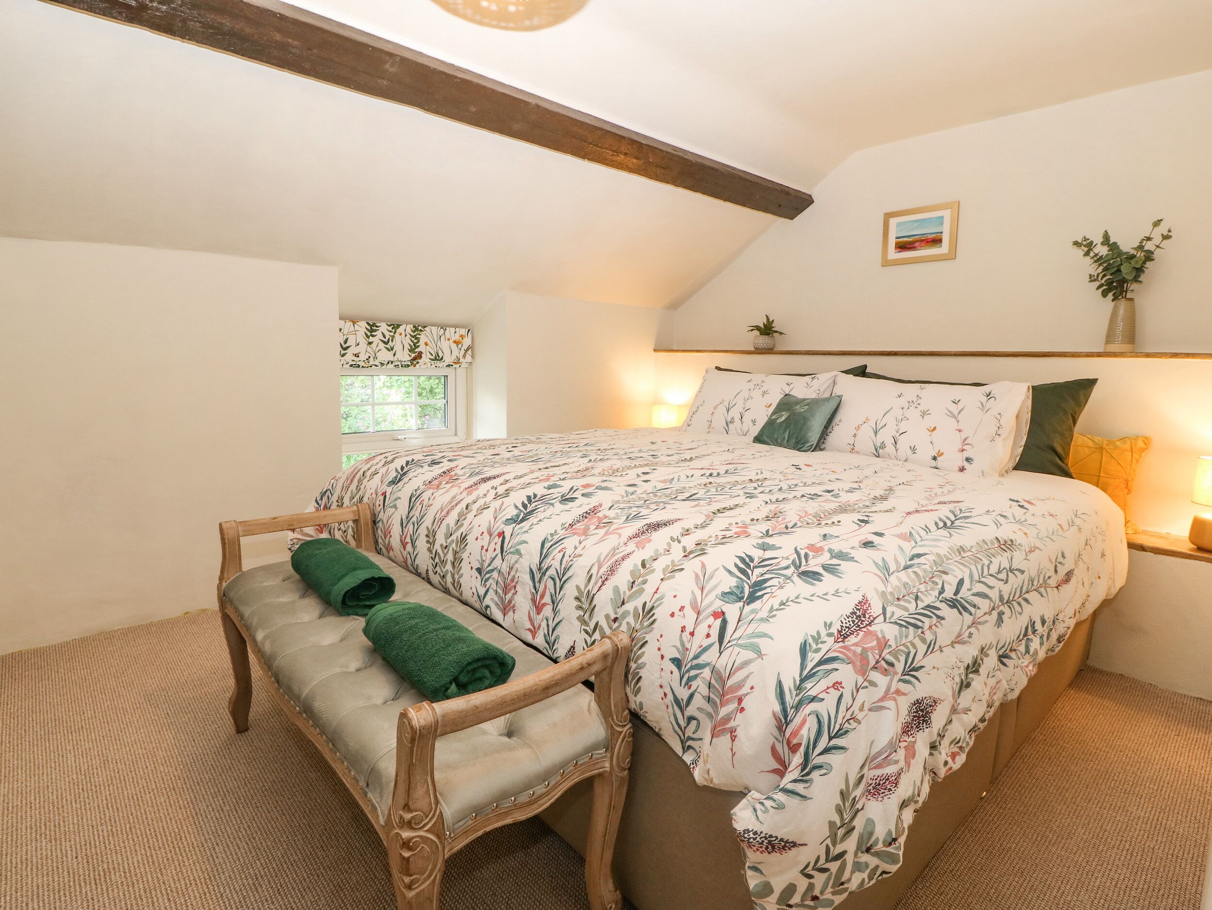A bedroom with a large bed, a wooden bench with green towels, a small window, and a wall-mounted plant.