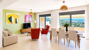 A spacious living area with a large window offering a view of the landscape, a television, and a dining area with chairs and a table. - Minimalist, new condo with beautiful sea and mountain views (Aegina)