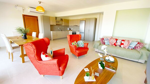 A modern apartment with a dining area, a living room with a sofa and armchairs, and a kitchen in the background.