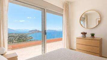 Une chambre avec un grand lit, une commode, un miroir rond et une vue sur un paysage côtier, visible à travers une porte-fenêtre coulissante.