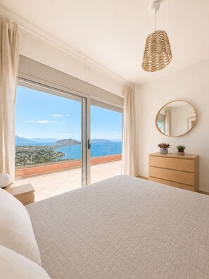 A bedroom with a large bed, a dresser, a round mirror, and a view of a coastal landscape through a sliding glass door. - Minimalist, new condo with beautiful sea and mountain views (Aegina)