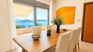 Een eetruimte met een houten tafel, witte stoelen, een potplant en uitzicht op zee door grote ramen.