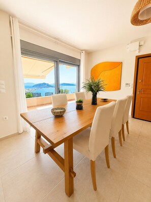 A dining area with a wooden table, white chairs, a potted plant, and a view of the sea through large windows. - Minimalist, new condo with beautiful sea and mountain views (Aegina)