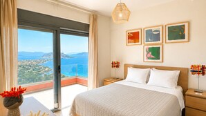 A bedroom with a large bed, a nightstand, a bedside table, a hanging light, and a view of the sea. - Minimalist, new condo with beautiful sea and mountain views (Aegina)