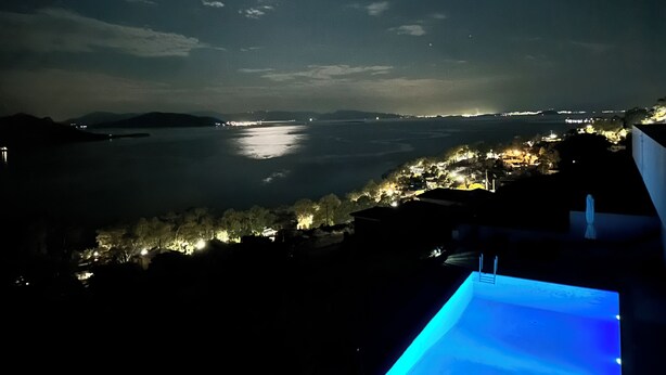 A nighttime view of a coastal area with a pool area and distant city lights. - Minimalist, new condo with beautiful sea and mountain views (Aegina)