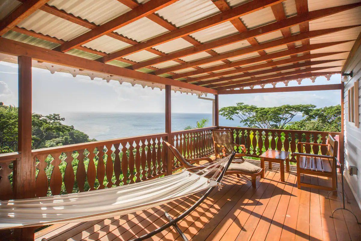 Terraza de madera con hamaca, mesa y sillas, con vista a un paisaje exuberante y el mar.