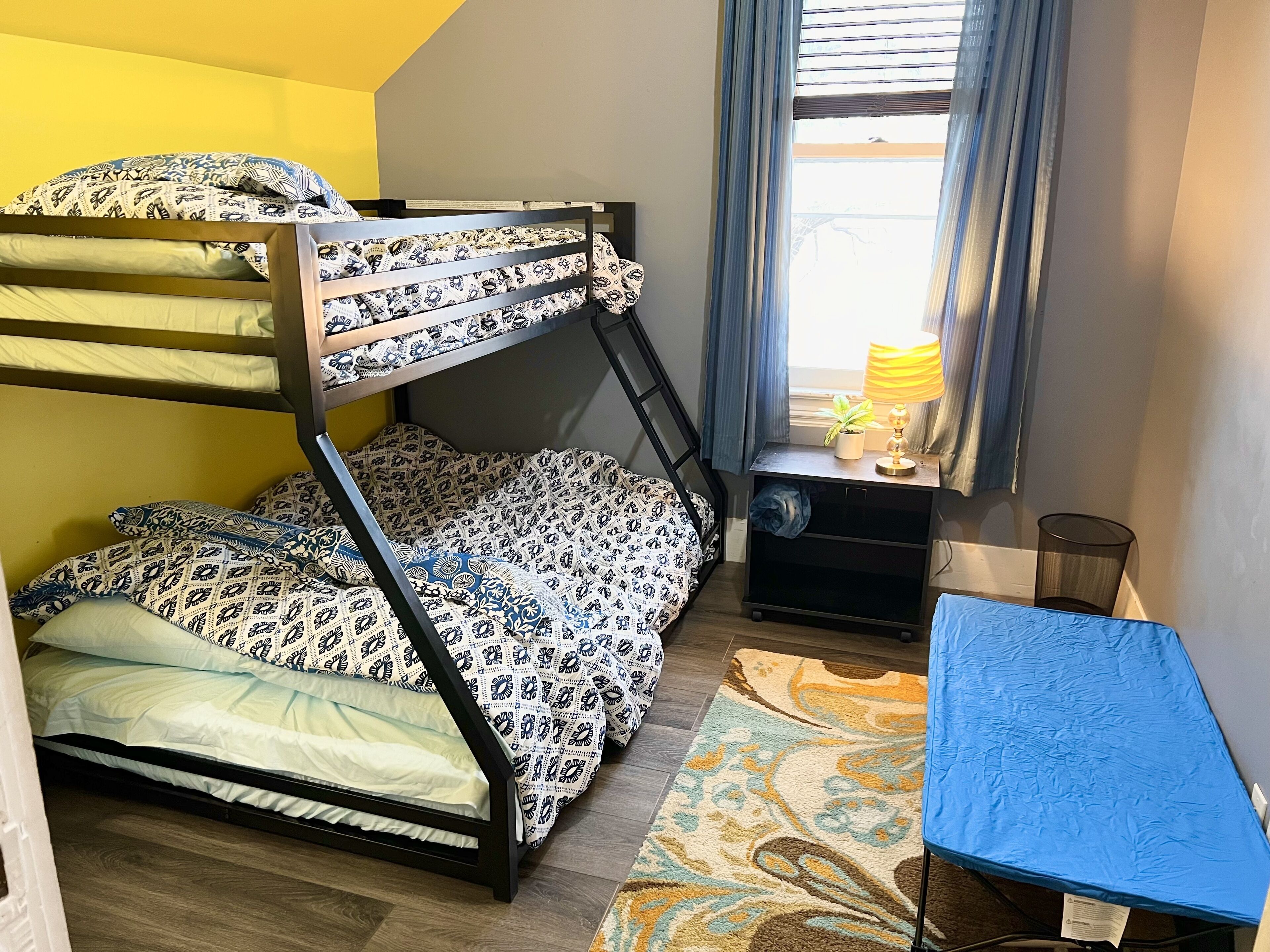 Una cama litera con ropa de cama estampada, un escritorio pequeño con lámpara y una ventana con cortinas.