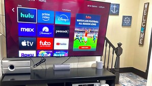 Un televisor TCL Roku sobre un mueble negro para centro de entretenimiento, con un control remoto y un dispositivo de transmisión.