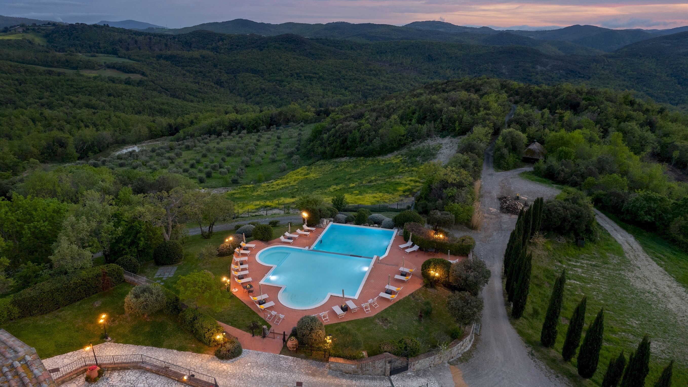 Vue aérienne d’un complexe hôtelier doté d’une piscine, entourée de chaises longues et bordée d’une allée pavée.