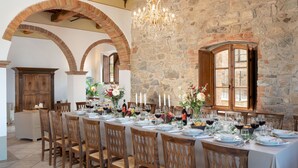 Uma longa mesa de jantar posta com cadeiras, velas e arranjos florais em um ambiente com paredes de pedra e vigas de madeira.
