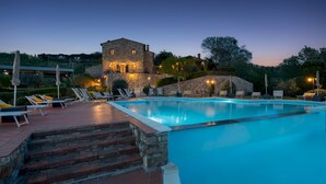 Uma piscina com espreguiçadeiras, um edifício de pedra com iluminação e uma escadaria que dá para a piscina.