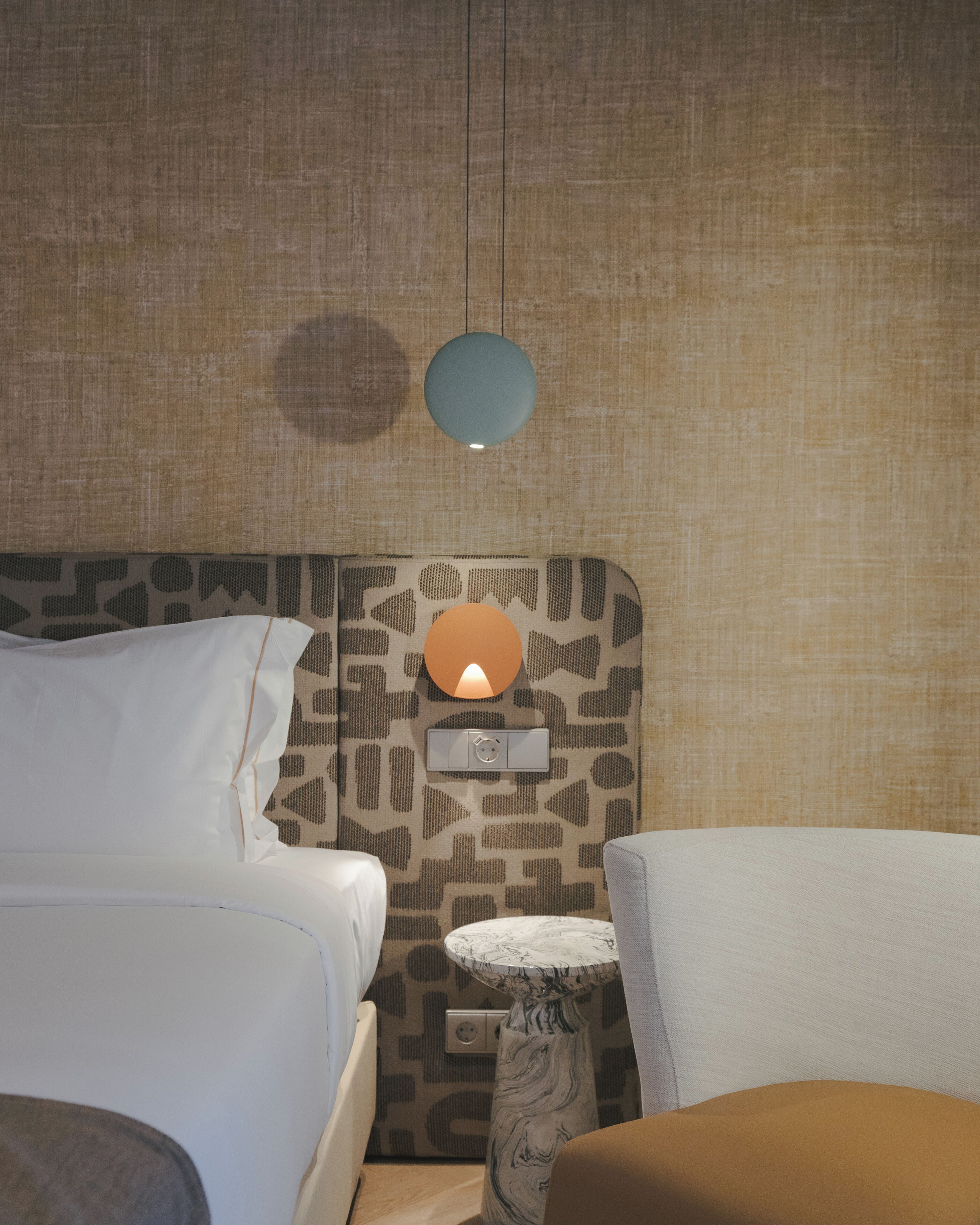 Una habitación de hotel con un cabecero estampado, una mesita de noche con un jarrón y una lámpara colgante.