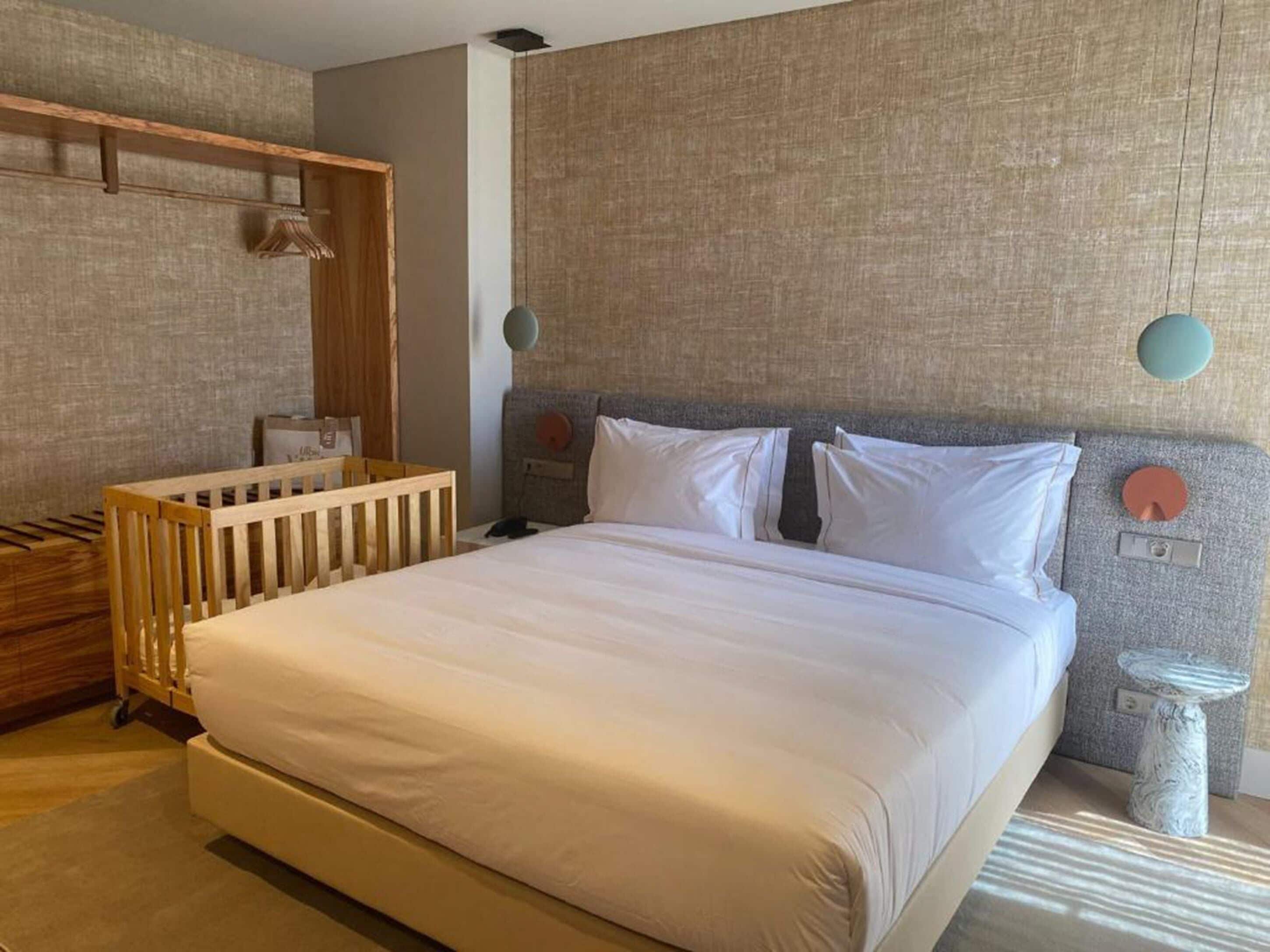 A bedroom with a large bed, a wooden crib, a bedside table, and a wall-mounted shelf.