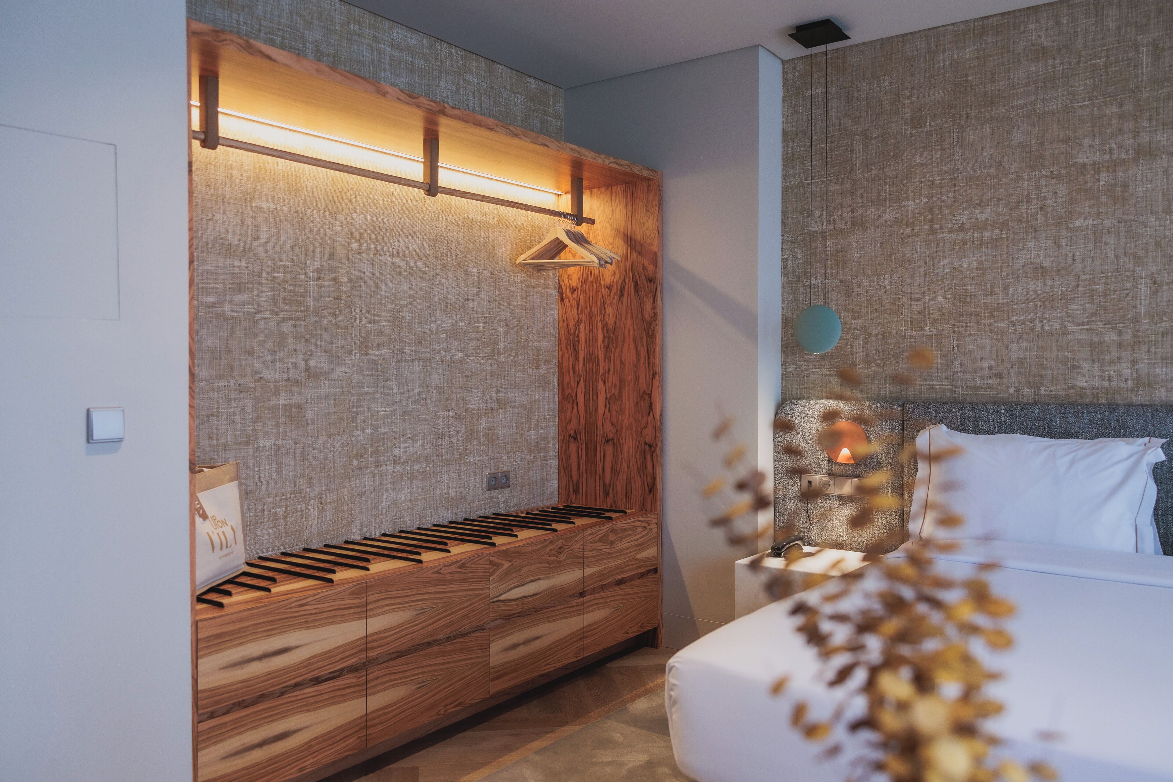 A modern bedroom with a wooden bench, a bedside table, and a hanging light fixture.