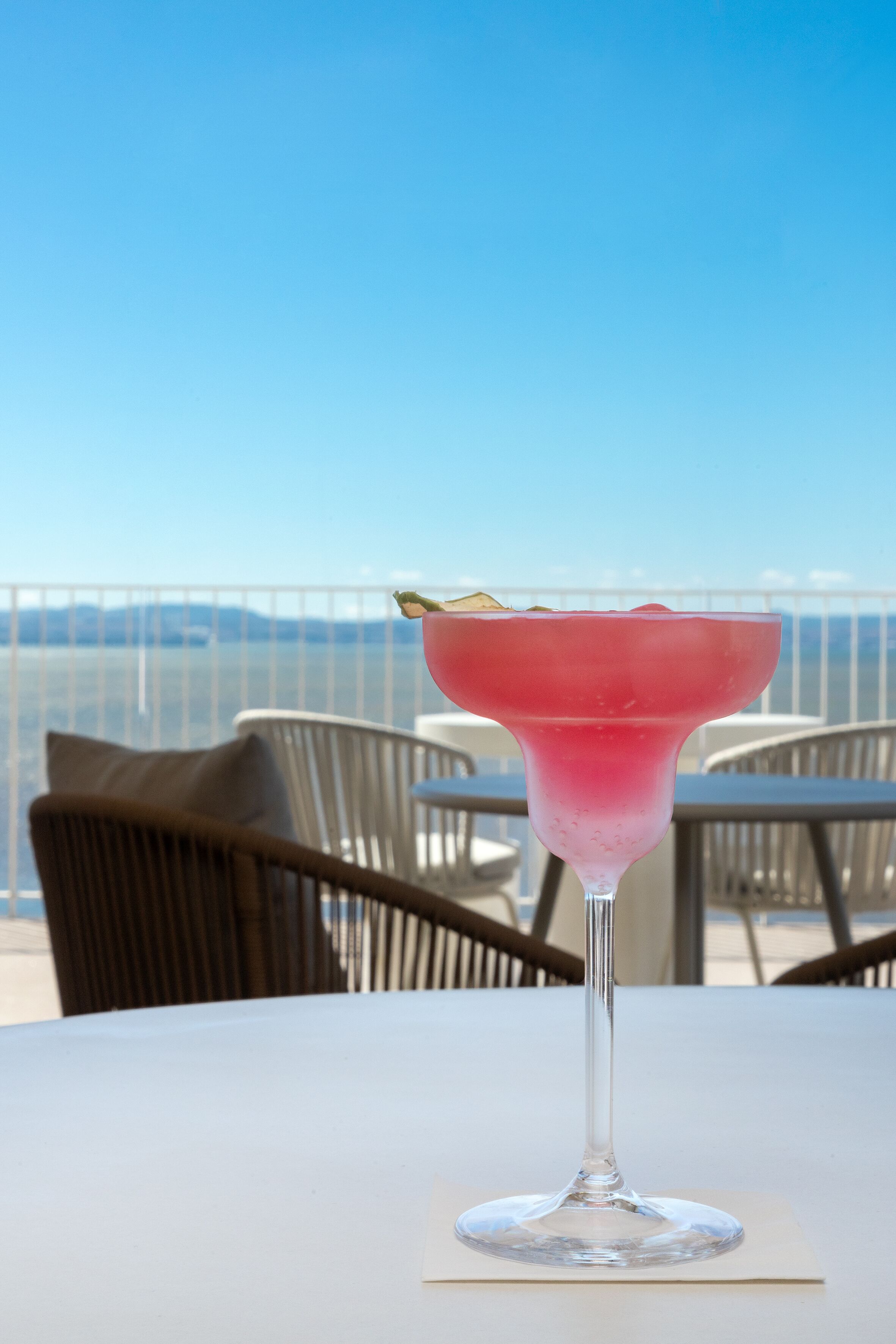 Un cóctel rosado sobre una mesa blanca, con vista al mar y montañas al fondo.