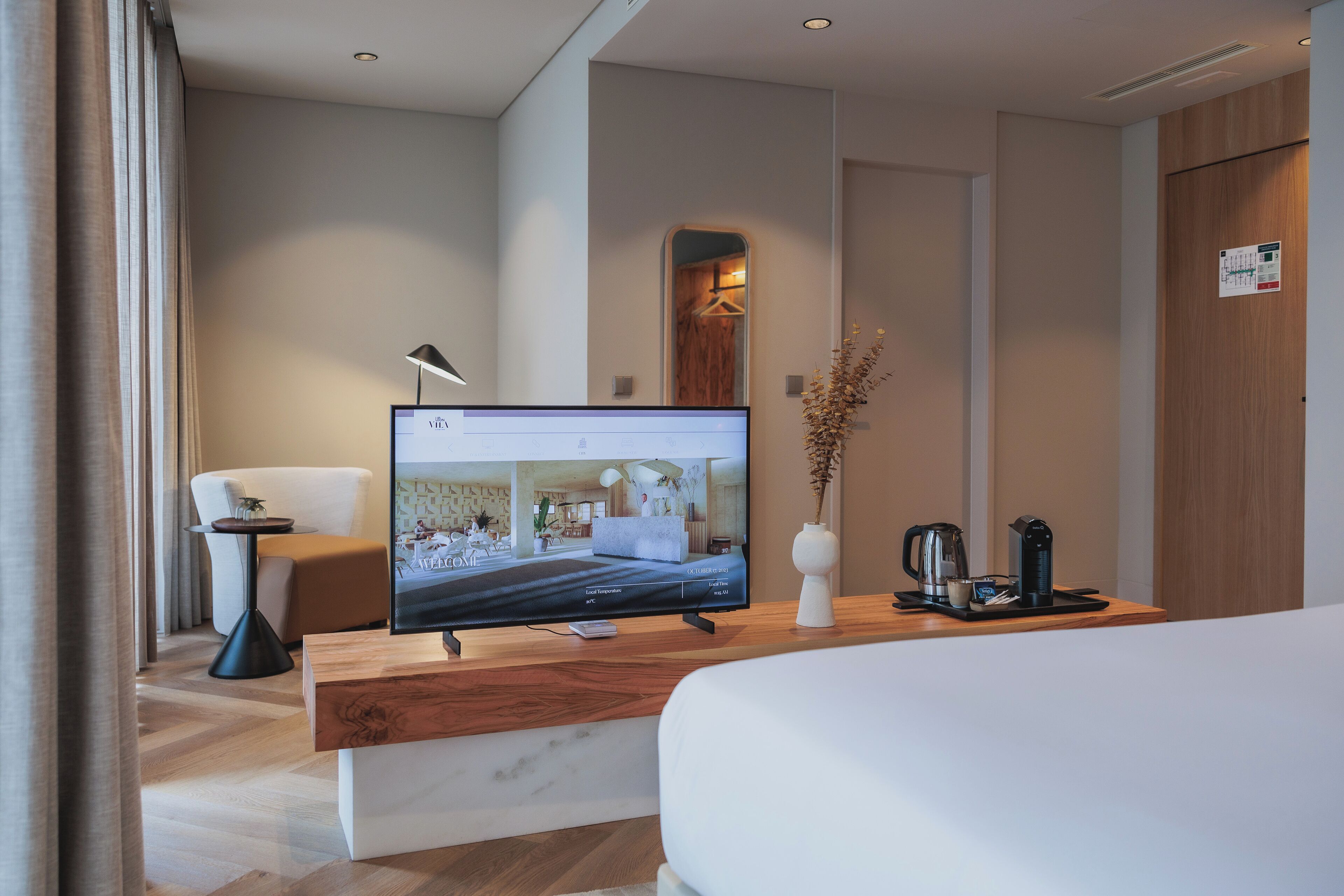 Quarto de hotel com uma TV de tela plana grande, uma escrivaninha de madeira, uma cama, uma cafeteira e um vaso com flores secas.