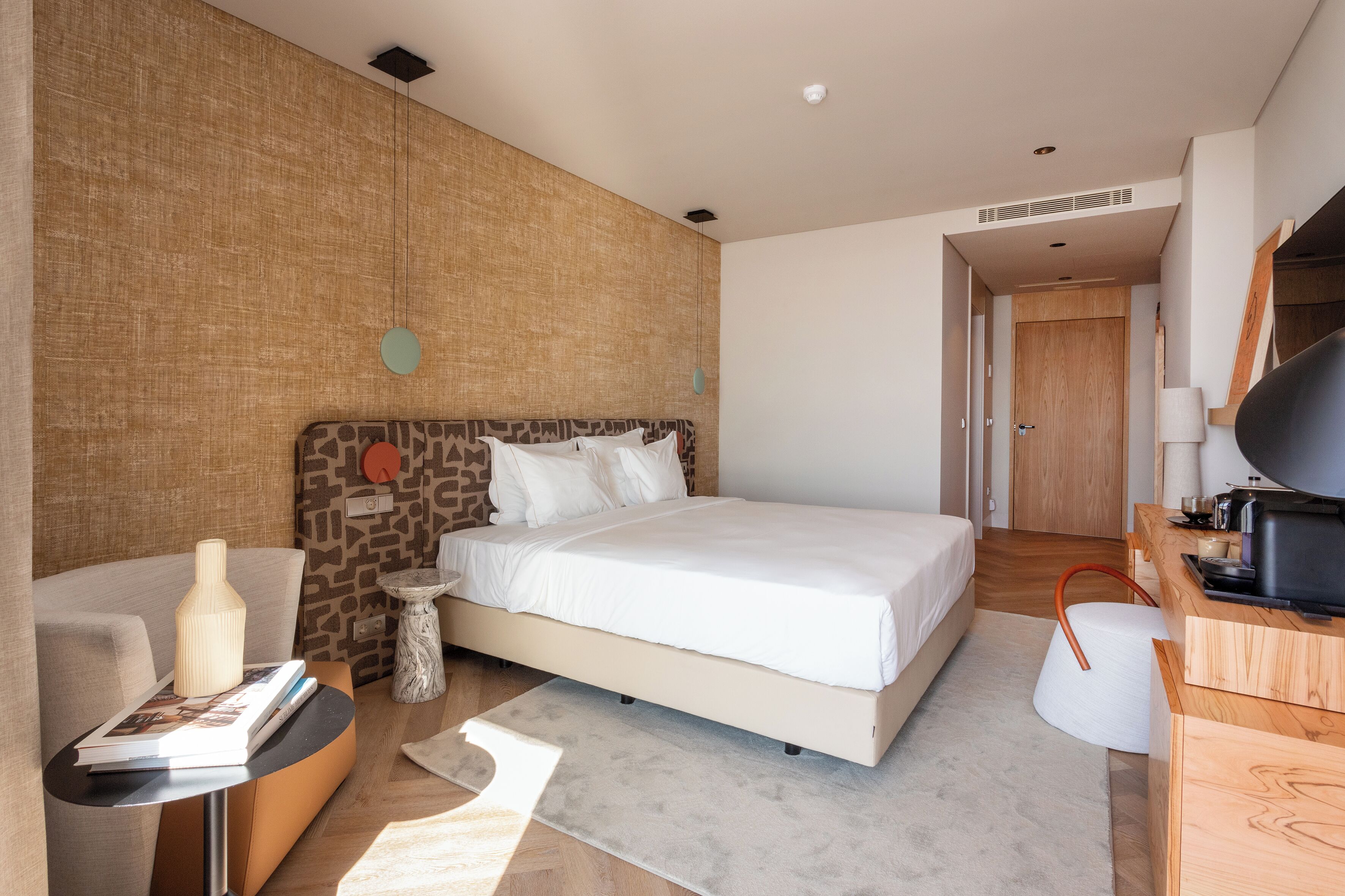Una habitación de hotel moderna con una cama grande, una mesita auxiliar pequeña, un sofá, un escritorio de madera y un televisor.