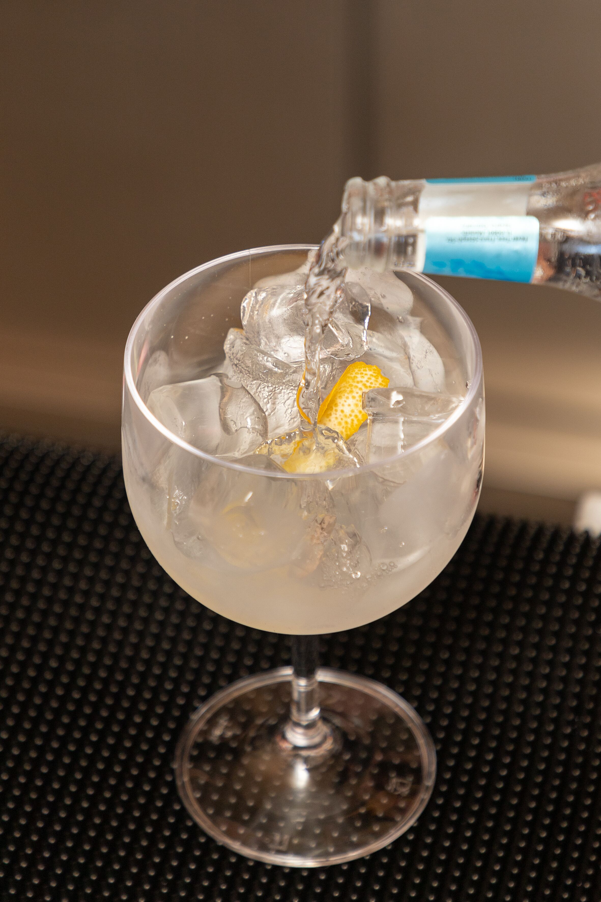 A clear glass with ice cubes and a lemon slice, being filled with a clear liquid from a bottle.