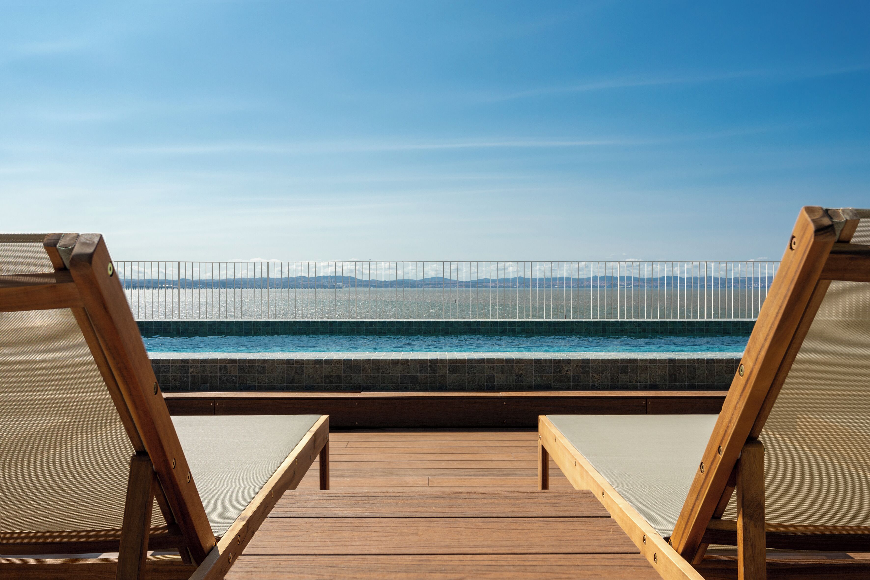 Una terraza de madera con dos sillones de descanso frente a una piscina y una vista despejada de montañas a lo lejos.