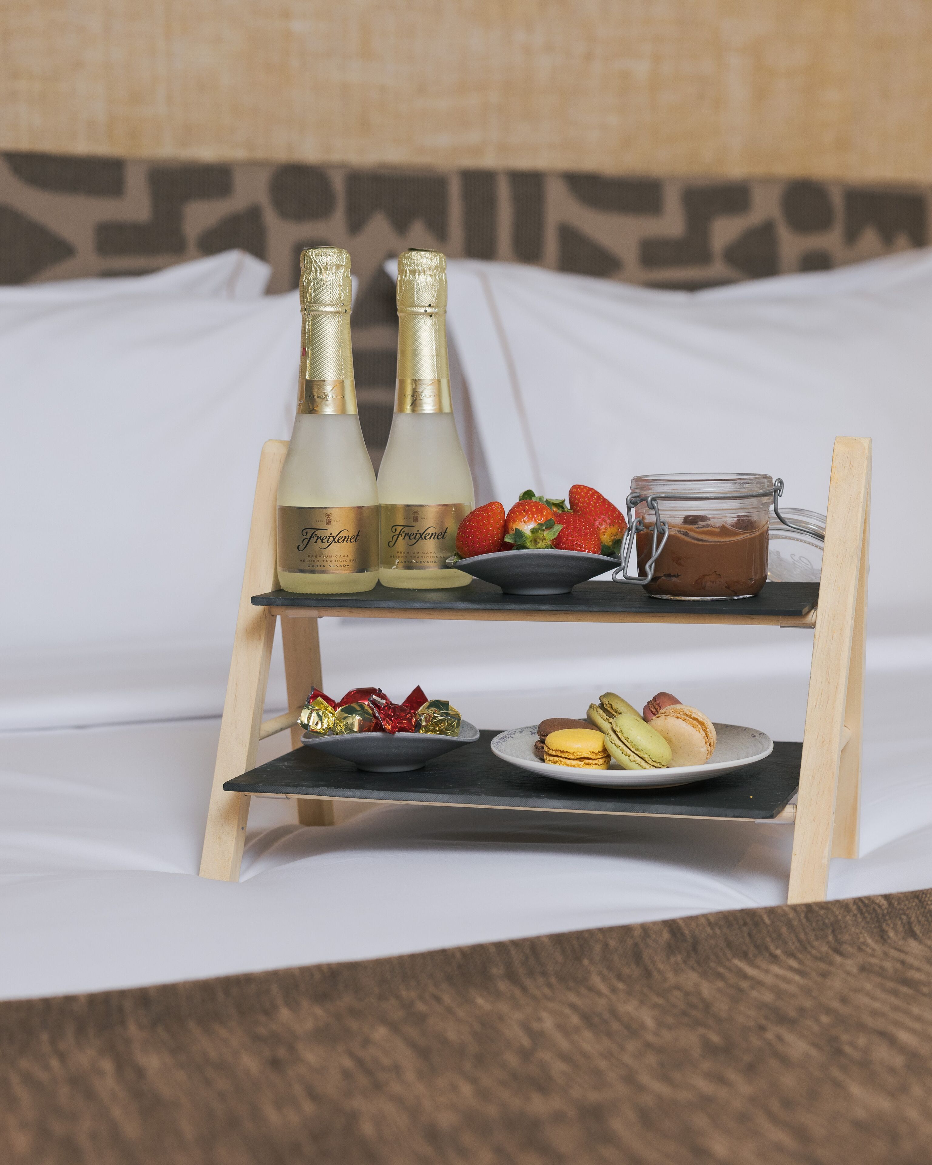 A tray with two bottles of Freixenet champagne, a jar of chocolate spread, strawberries, macarons, and foil-wrapped chocolates on a bed.
