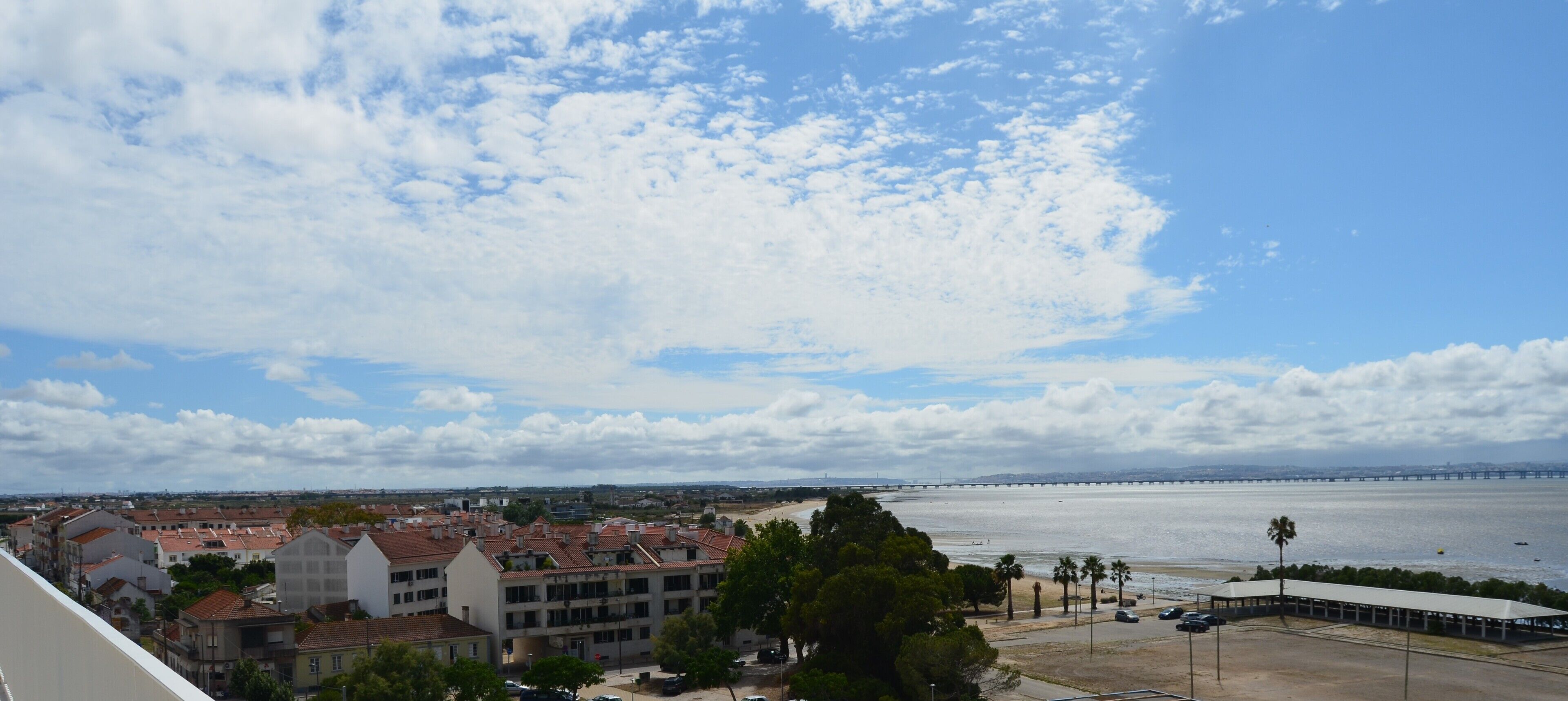 A coastal cityscape with a bridge in the distance, buildings with red-tiled roofs, and a clear sky.
