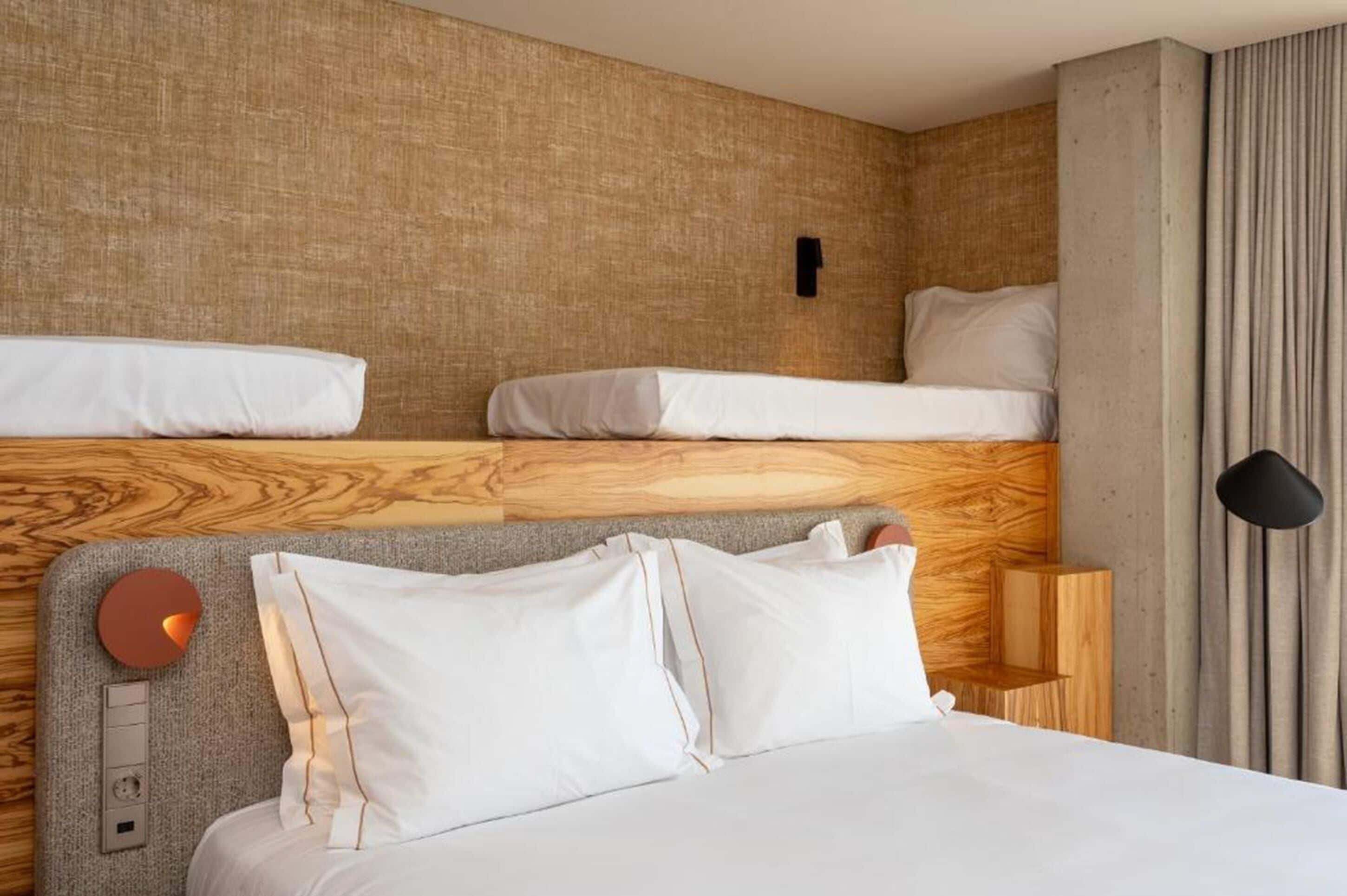 Quarto de hotel com uma cama beliche, uma mesa de cabeceira com abajur e um painel de controle fixado na parede.
