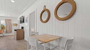 Ein modernes Zimmer mit Holztisch und weißen Stühlen, einem Fernseher und zwei gewobenen Wandspiegeln.