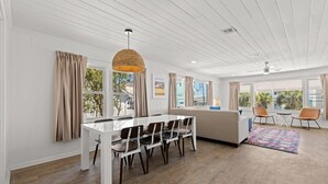 Ein geräumiges Zimmer mit weißer Decke, einem Esstisch mit Stühlen, einer Couch und einem großen Fenster mit Vorhängen.