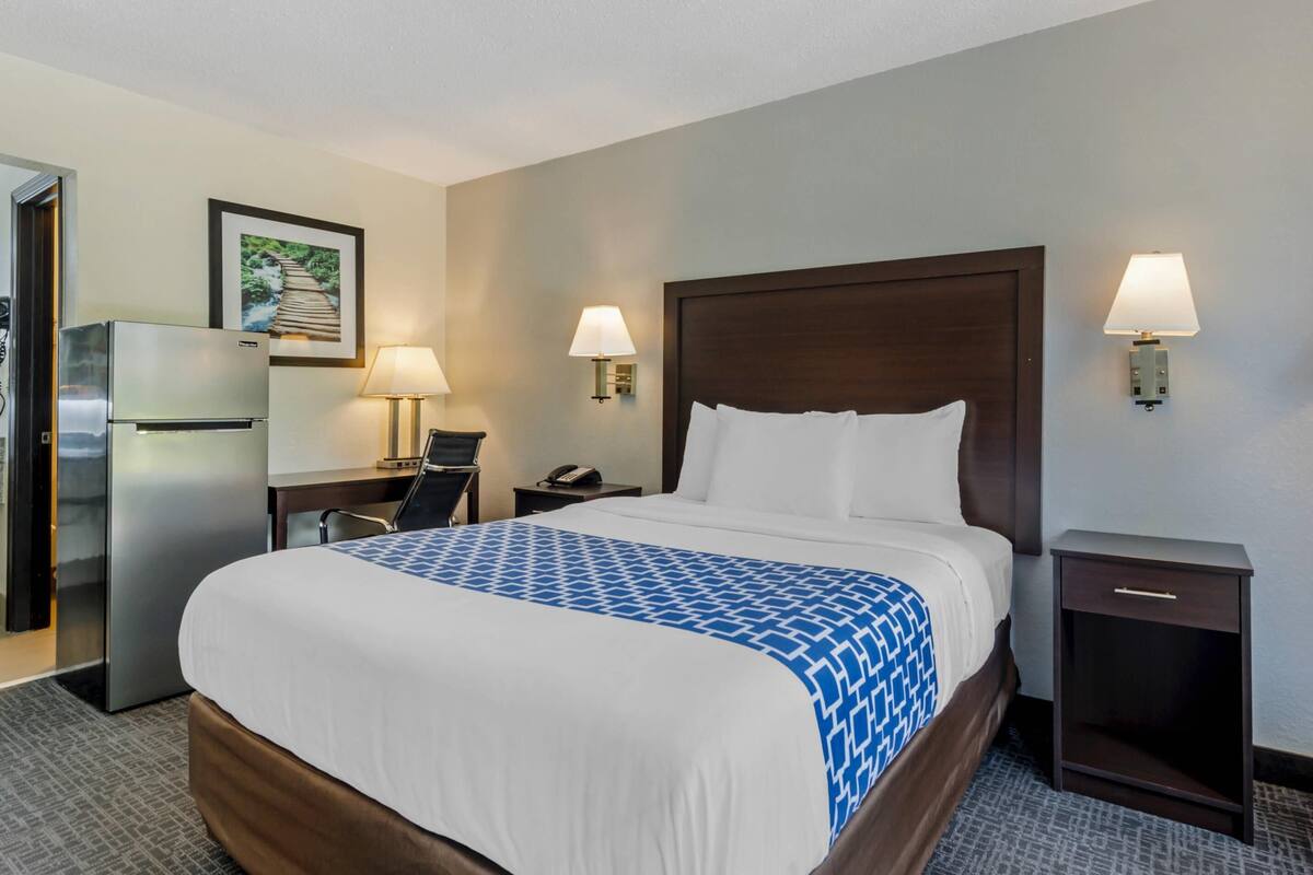 A hotel room with a large bed, two bedside lamps, a desk with a lamp, a refrigerator, and a wall-mounted picture.