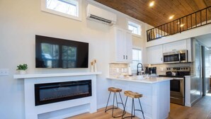 A modern kitchen with white cabinetry, a large island with stools, a flat-screen TV above the fireplace, and a stainless steel oven.