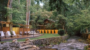 A wooden cabin with a deck and lounge chairs, surrounded by a stone wall and a stream. - Sage Cabin by Creekside Hideaway (Gatlinburg)