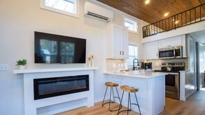 A modern kitchen with white cabinetry, a built-in TV, a fireplace, and a bar area with stools. - Sage Cabin by Creekside Hideaway (Gatlinburg)