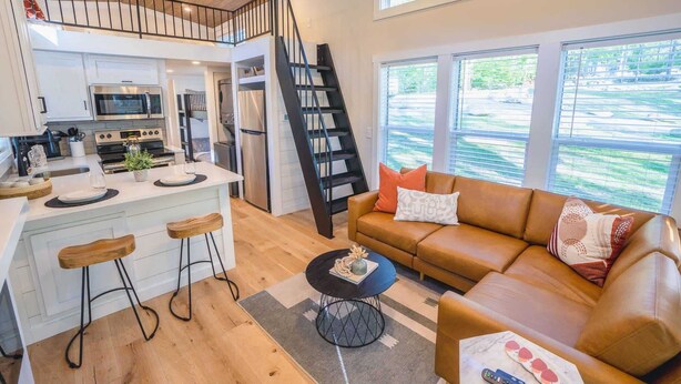 A modern kitchen with a central island, a staircase, a living area with a leather sofa, and large windows.
