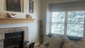 Una sala de estar acogedora con chimenea, un sofá gris y un ventanal con vistas a árboles.