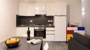 A compact kitchen with white cabinets, a black countertop, a stove, and an oven. There is a dining table with a bowl of fruit and a wine bottle. - Guelfa 14 - Two Bedroom Apartment, Sleeps 4 (Firenze)