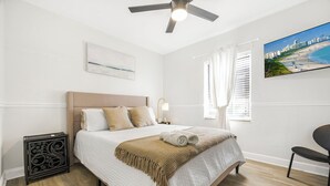 Quarto com cama, ventoinha no teto, mesa de cabeceira, cadeira, janela com cortinas e um quadro na parede.