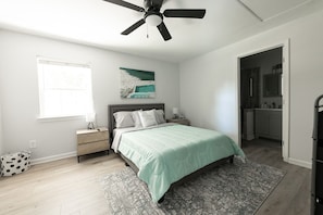 Quarto com cama, mesa de cabeceira, ventilador de teto, janela e banheiro visível por uma porta aberta.