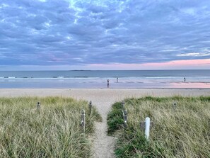 Camí de platja que condueix a la riba, amb dunes herboses i un cel serè.