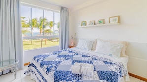 Ein Hotelzimmer mit einem großen Bett, einem Glastisch, Blick auf Palmen und Meer sowie einem Regal mit gerahmten Bildern.