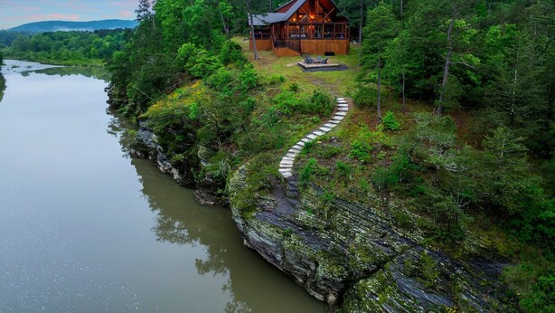 Une cabane en rondins avec un sentier de pierres menant à une terrasse, entourée d’une forêt dense et d’un lac.