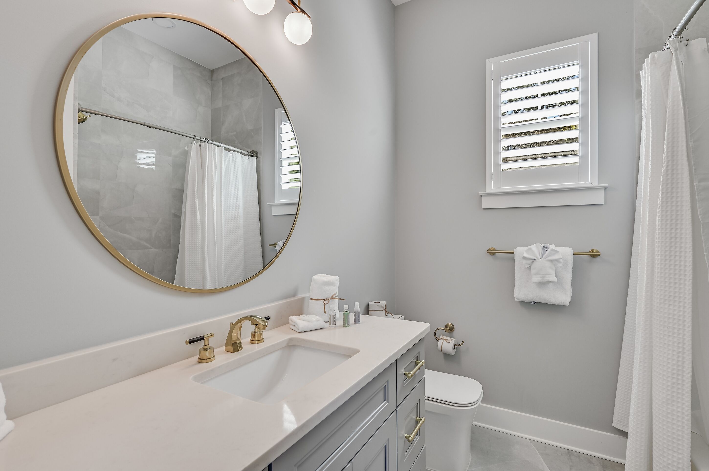 A bathroom with a round mirror, a sink, a toilet, and a window with blinds.