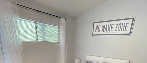 A bedroom with a bed, a black sofa, a nightstand with a lamp, and a sign that reads "NO WAKE ZONE".