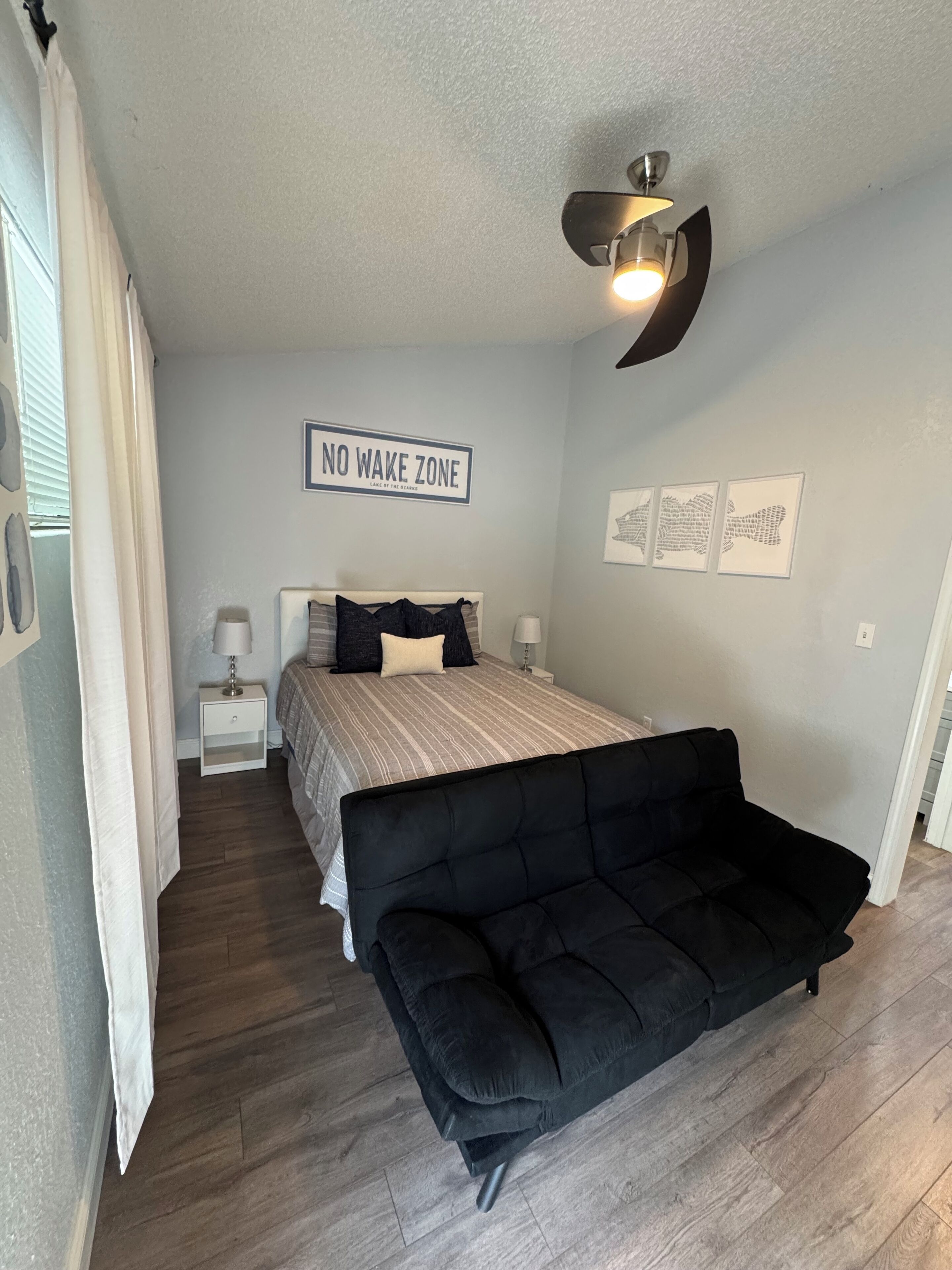 Een slaapkamer met een bed, een zwarte bank, een plafondventilator, een bordje aan de muur met de tekst 'NO WAKE ZONE' en lijstjes met kunstwerken aan de wand.