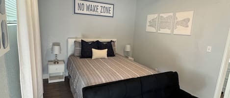 Een slaapkamer met een bed, een zwarte bank, een plafondventilator, een bordje aan de muur met de tekst 'NO WAKE ZONE' en lijstjes met kunstwerken aan de wand.