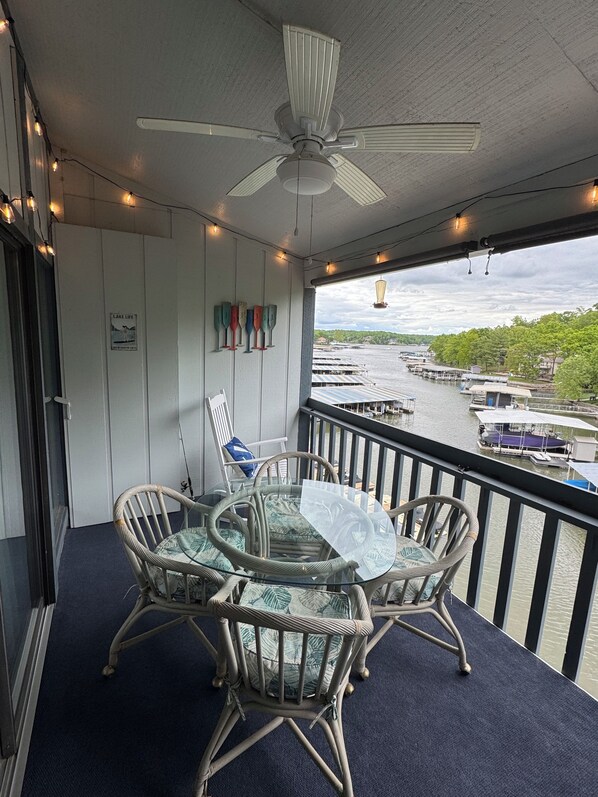 A balcony with wicker chairs and a glass table, overlooking a marina with boats and a lake.
