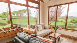 Ein gemütliches Wohnzimmer mit einer Couch, einem Couchtisch und einem Kicker. Große Fenster bieten Blick auf eine grüne Landschaft.