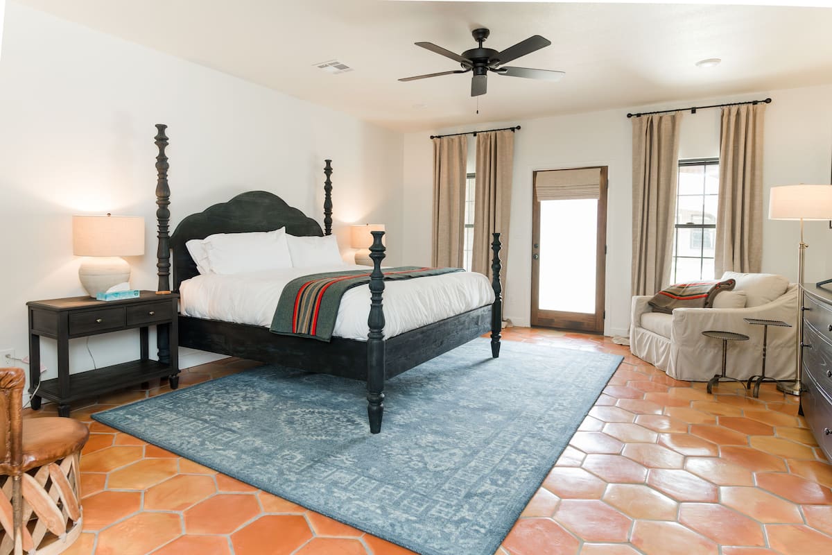 A bedroom with a four-poster bed, a ceiling fan, a nightstand with a lamp, a chair, a dresser, and a window with curtains.