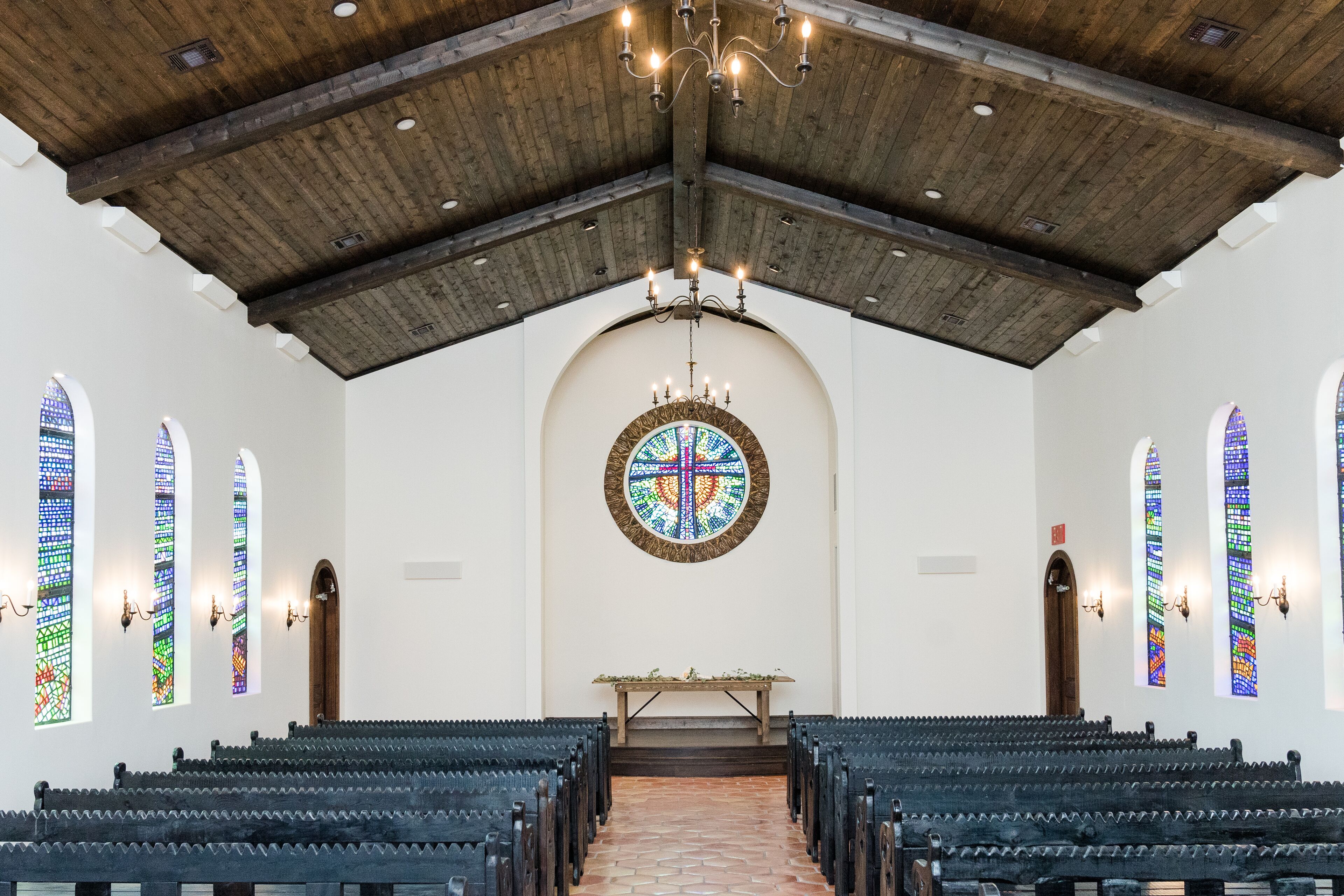 Ein Kircheninneres mit Kirchenbänken, einer Holzbalkendecke, Buntglasfenstern und einem zentralen Altar.