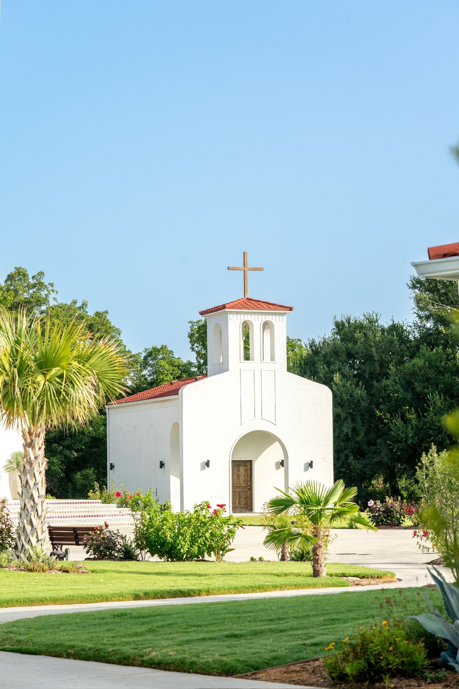 Una iglesia blanca con tejado rojo y una cruz en la parte superior, rodeada de vegetación y un césped cuidado.