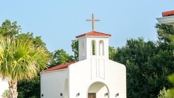 Una iglesia blanca con tejado rojo y una cruz en la parte superior, rodeada de vegetación y un césped cuidado.